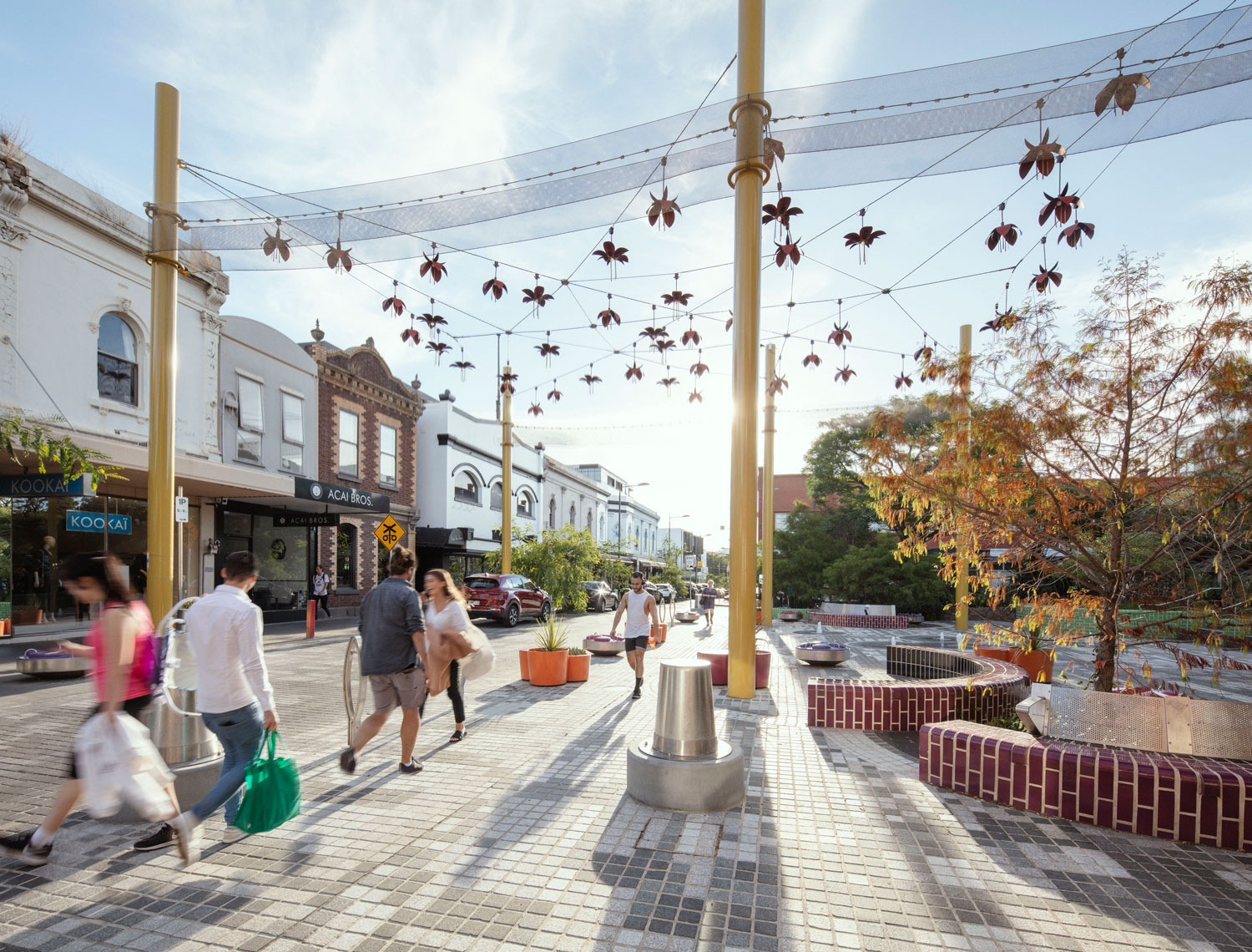 A photo of a paved pedestrian area. There are people waking around, trees and shops on the perimeter and an aerial artwork hanging above.