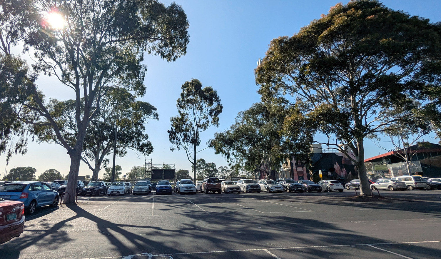Now: A wide view of the Russell Street east car park. An open air car park, looking towards City Oval. There are large trees in the mid ground.