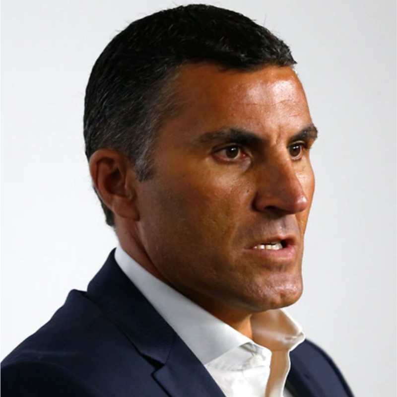 A man is in 3/4 profile to the camera. He has brown eyes, dark hair and tanned skin. He wears a blue suit jacket and white shirt.