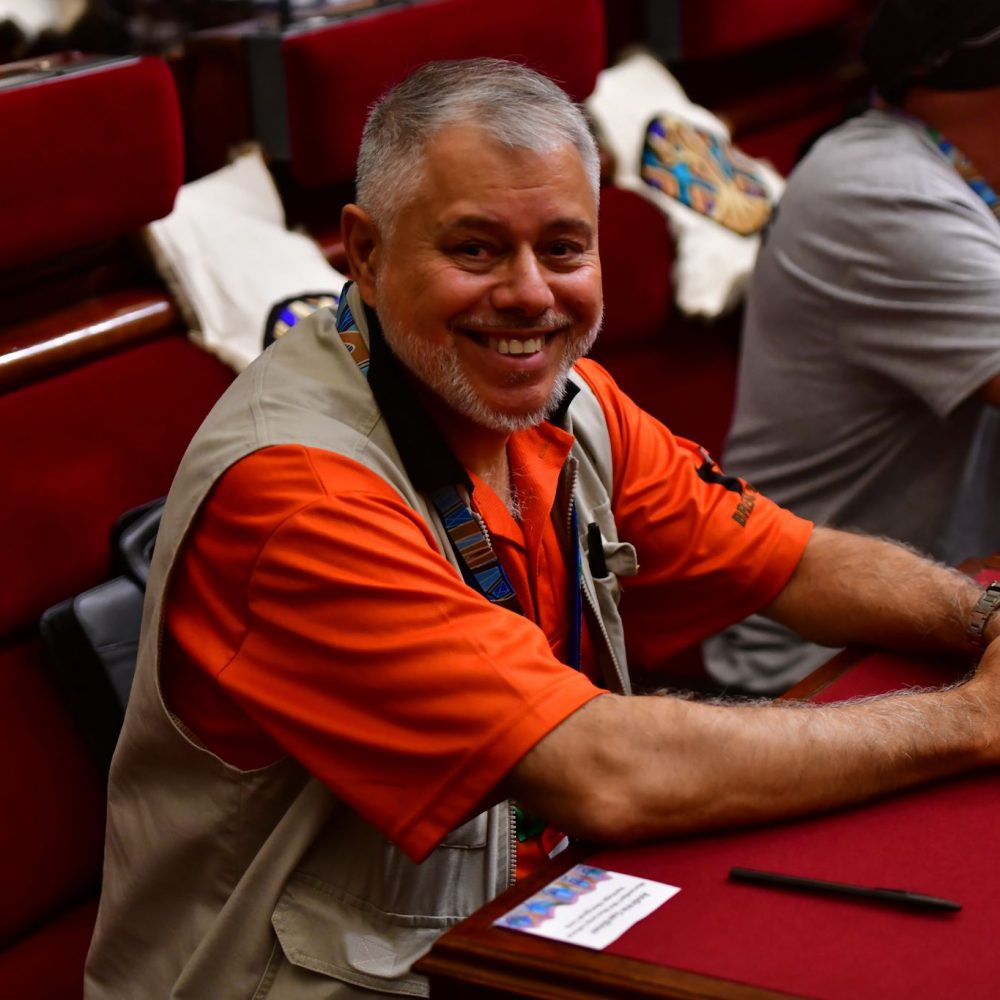 Uncle Andrew Gardiner is a bearded man smiling at the camera. He wears an orange t shirt with a beige vest.