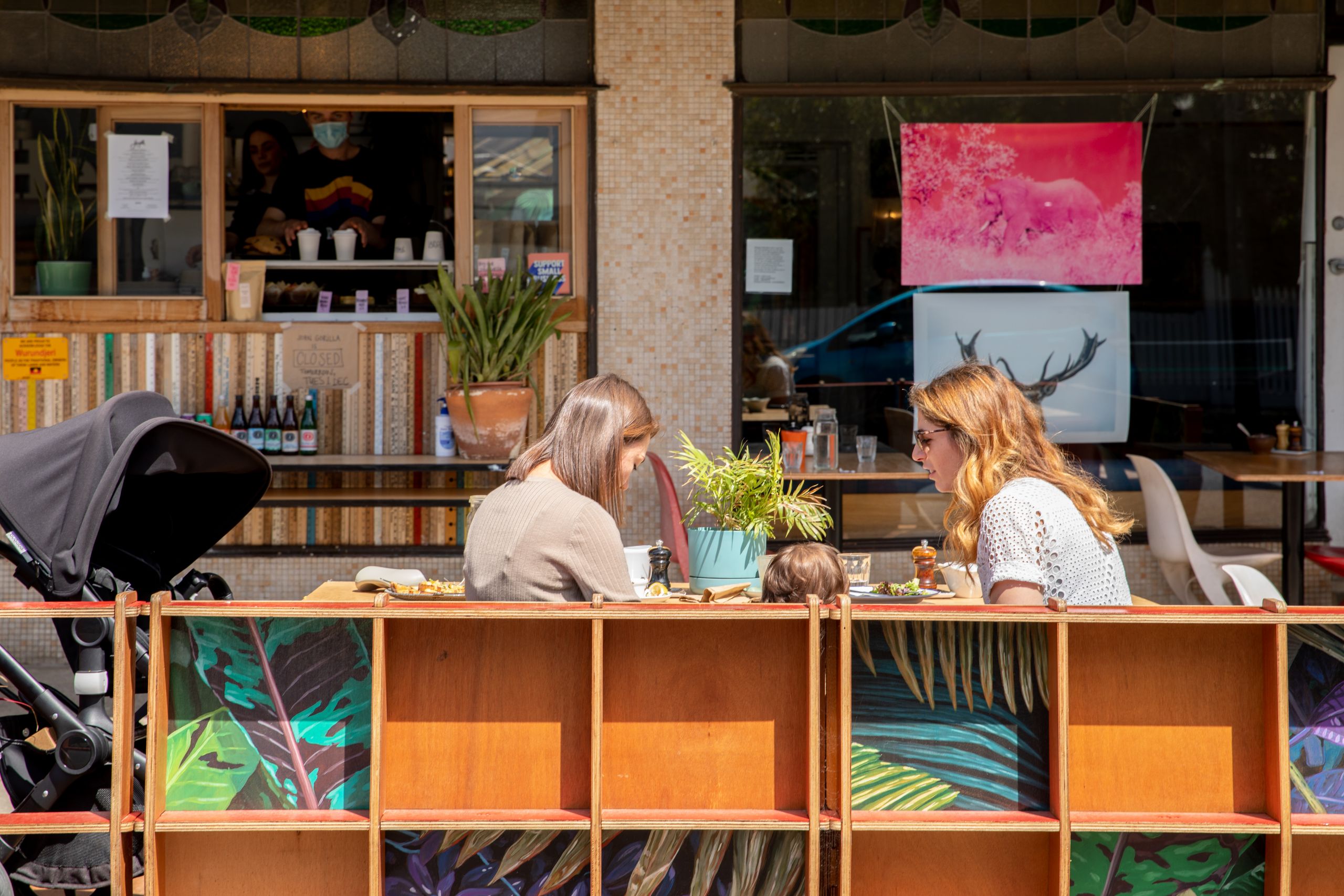 2 women sit opposite each other, with a child in between them, on the footpath outside a cafe. There is a wooden structure to protect them from the street.