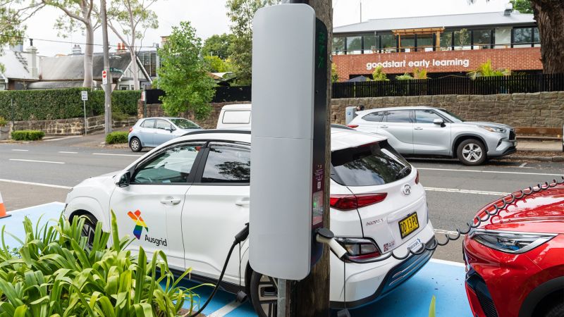 A close up of a pole-mounted EV charger. It is a slim grey box attached to a wooden power pole. In the background there is a car parked on the street, near the pole with a spiral cord running from the charger to the car.