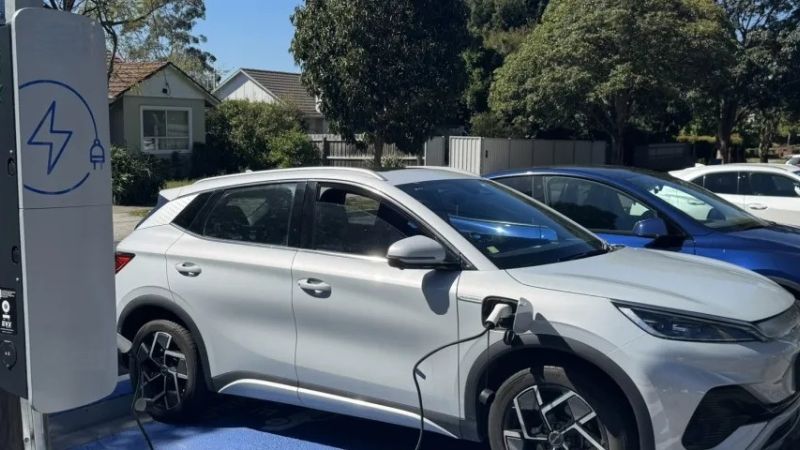A car parked in a car park space with a charging cable running from the car to a small grey box mounted on a wooden pole. 