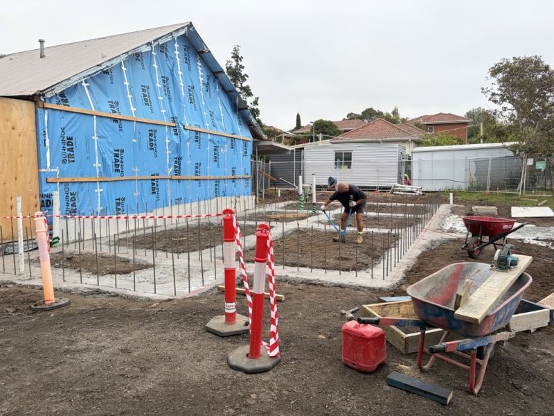 The side of a building under construction. There is blue tarp covering where the building is open to the elements. There are two people working on the ground next to the building where there are concrete foundations for garden beds.