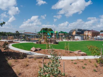 A brand new park on a sunny day. there are small seedlings planted in mulch and a wide expanse of lawn with a playground in the distance.