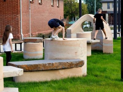 3 children are climbing and playing on an outdoor scultural work. The scultpure is made of a light coloured stone that is shaped into cylanders, seats and other geometric shapes. It is placed on the grass and has a look of lightness despite the heavy materials.