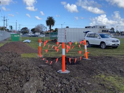 A hole in the muddy ground surrounded by orange hazard tape with a white car and shipping container in the background.