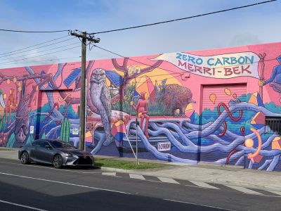 A mural on the outside wall of a 1 storey building. the artwork depicts small pixie-like people sitting among different Australian animals and plants. There is an old car with tree roots growing out of it. The colours are like a sunset.