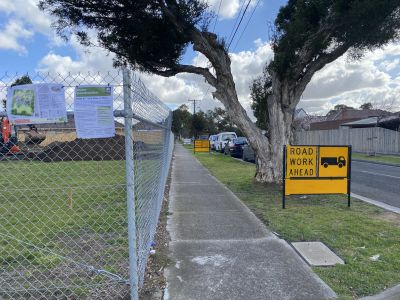 A footpath with a roadwork ahead sign on the nature strip on the right and chain fencing around a construction site on the left.