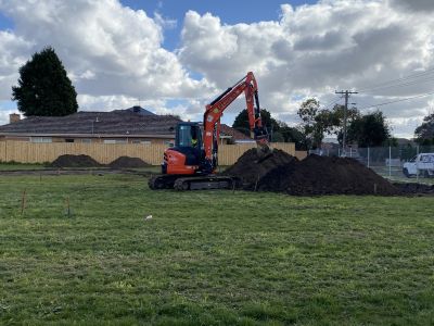 An orange digger is piling up mounds of dirt in an open green space, with a house behind a fence in the background
